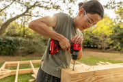 A person wearing safety goggles, a mask, and ear protection uses a red CRAFTSMAN cordless drill to cut a hole in a ceiling above a window.