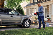 Photo of person using CRAFTSMAN CMCPW350B cordless power cleaner to wash a gray SUV in a driveway in front of a house.