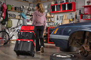 A person is pulling a stack of black and red rolling toolboxes in a garage workshop. The garage contains a bicycle, a blue car with the wheel removed, shelves with various tools and supplies, and a workbench.