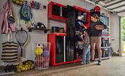 A garage scene featuring CRAFTSMAN red and black storage cabinets and shelving filled with sports equipment, toys, and miscellaneous items. Two people, an adult and a child, are organizing items on the shelves. There are various sports balls in a white wire basket, a tennis racket, bags, and shoes, with a wall-mounted shelf and hooks holding more equipment.