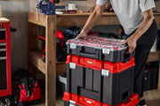 A person is stacking three CRAFTSMAN modular tool storage boxes with black bodies and red accents in a workshop setting. The top box has a clear lid, and the boxes are positioned on a wooden workbench with tools and a red tool bag visible nearby.