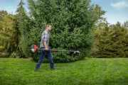 Photo of a person carrying a Craftsman CMXGTAMDSS30 string trimmer across a grassy lawn, with evergreen trees in the background.